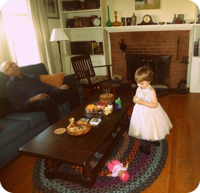 Toddler in a fancy dress playing in a living room, an older man on the couch laughing