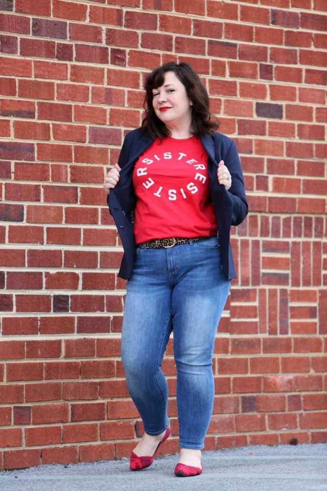 Woman in front of a brick wall wearing a t-shirt that says Resister Sister on it. She is wearing a navy double-breasted blazer over it and skinny jeans on the bottom half.