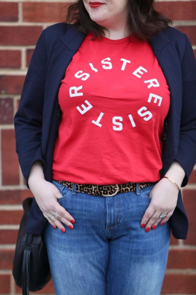 Woman in front of a brick wall wearing a t-shirt that says Resister Sister on it. She is wearing a navy double-breasted blazer over it and skinny jeans on the bottom half.