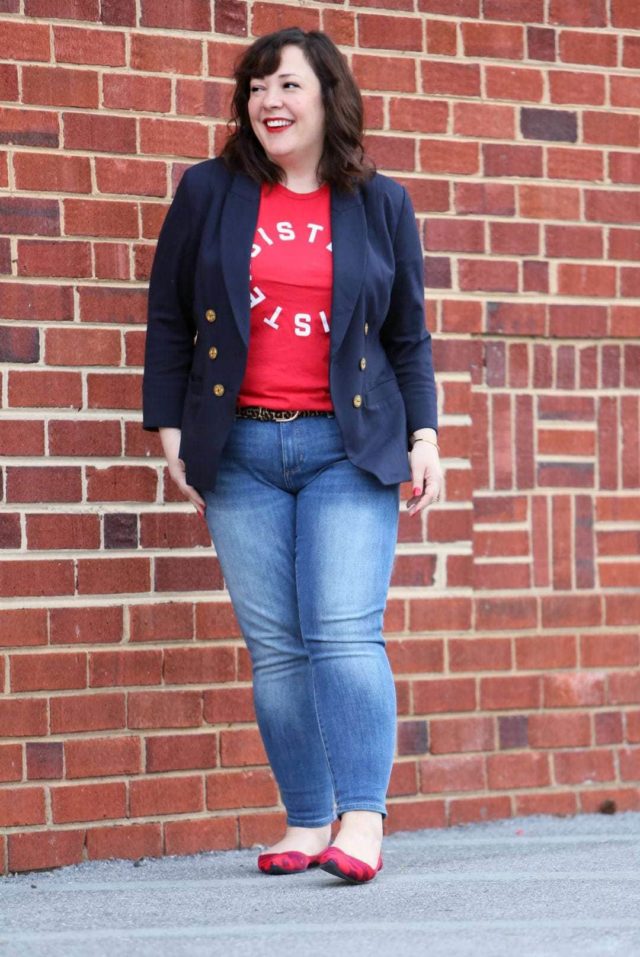 Woman in front of a brick wall wearing a t-shirt that says Resister Sister on it. She is wearing a navy double-breasted blazer over it and skinny jeans on the bottom half.