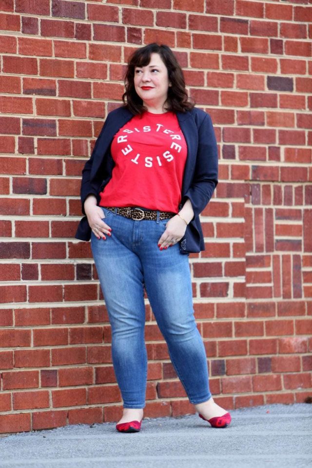 Woman in front of a brick wall wearing a t-shirt that says Resister Sister on it. She is wearing a navy double-breasted blazer over it and skinny jeans on the bottom half.