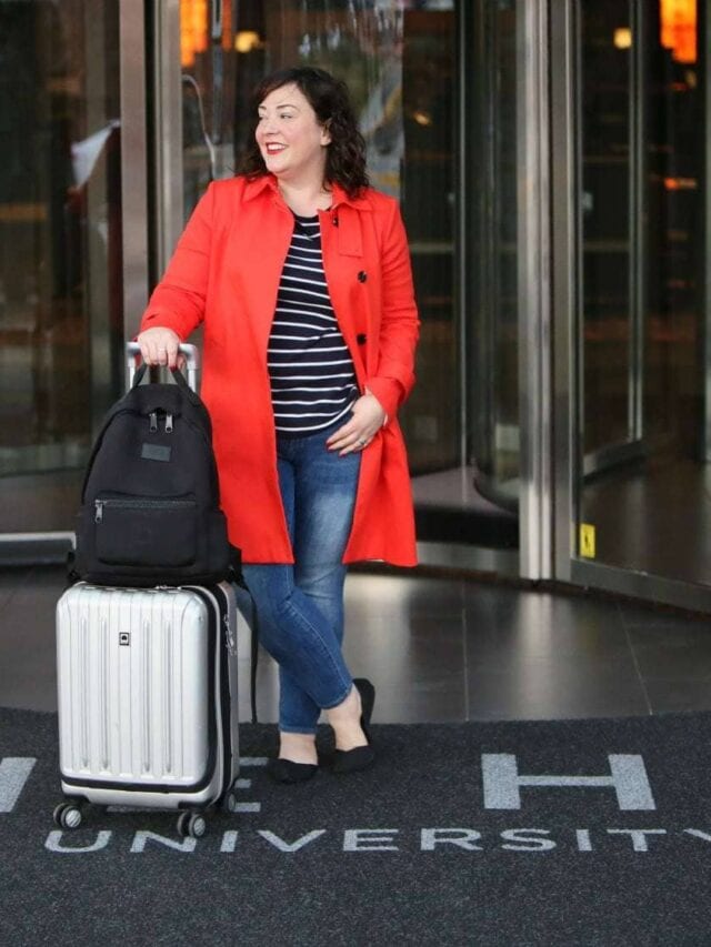 woman in a rd trench coat walking out of a hotel pushing a silver rolling suitcase with a black backpack on top of the suitcase