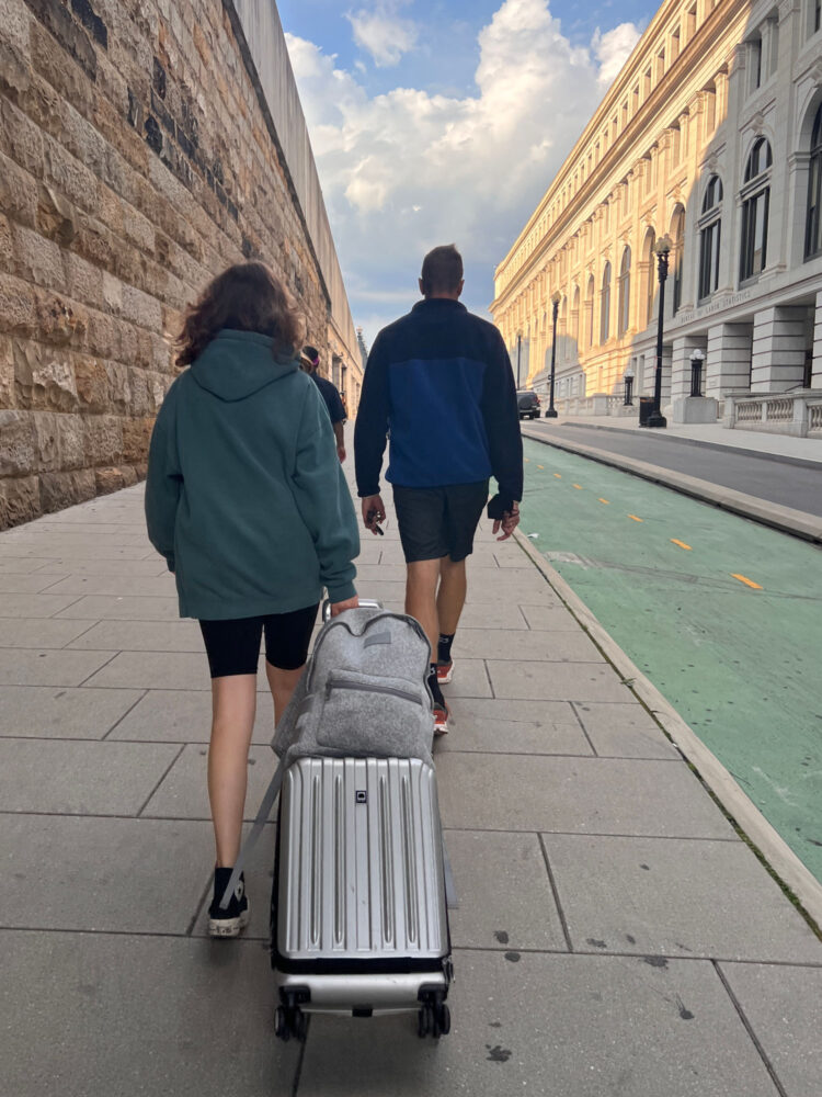 A teen and her father walking into a train station; the teen is pulling a rolling suitcase with a gray Dagne Dover Dakota backpack on top secured by its luggage sleeve. Dagne Dover Dakota backpack review by Wardrobe Oxygen