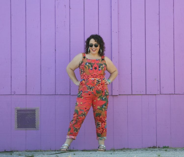 Nooworks Overalls Review 1 woman with dark brown shoulder length curly hair standing in front of a purple wall. She is wearing cotton overalls that fit more like a jumpsuit. They are an orange print with tigers all over them. She has styled the overalls with silver hoops, silver Birkenstock Arizona sandals, and a silver and turquoise cuff bracelet.