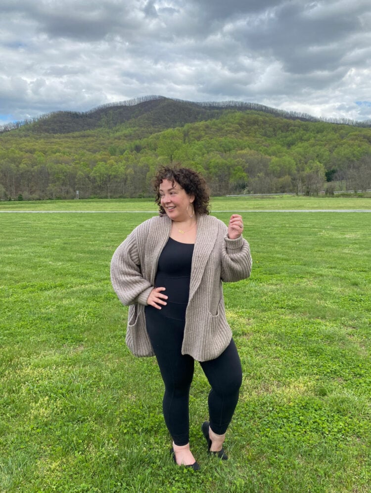 Alison Gary of Wardrobe Oxygen is standing in a field at a brewery in Berkeley Springs, WV. She is wearing a black Universal Standard next to Naked bodysuit with the M.M.LaFleur Snyder Jacket belted sweater in taupe.