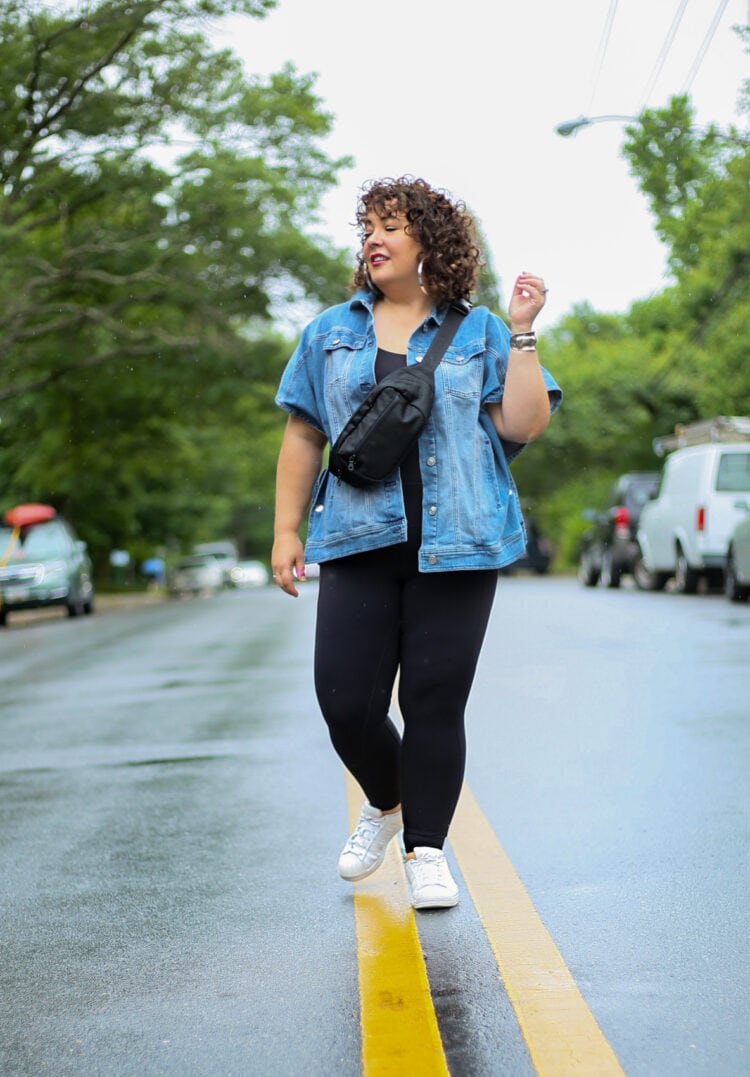 Alison standing in the middle of a street. She is wearing a hip length sleeveless denim jacket and black leggings. She has on white Adidas Superstar sneakers and has a black belt bag across her body. Alison is sharing How to Style the Universal Standard Next to Naked Bodysuit
