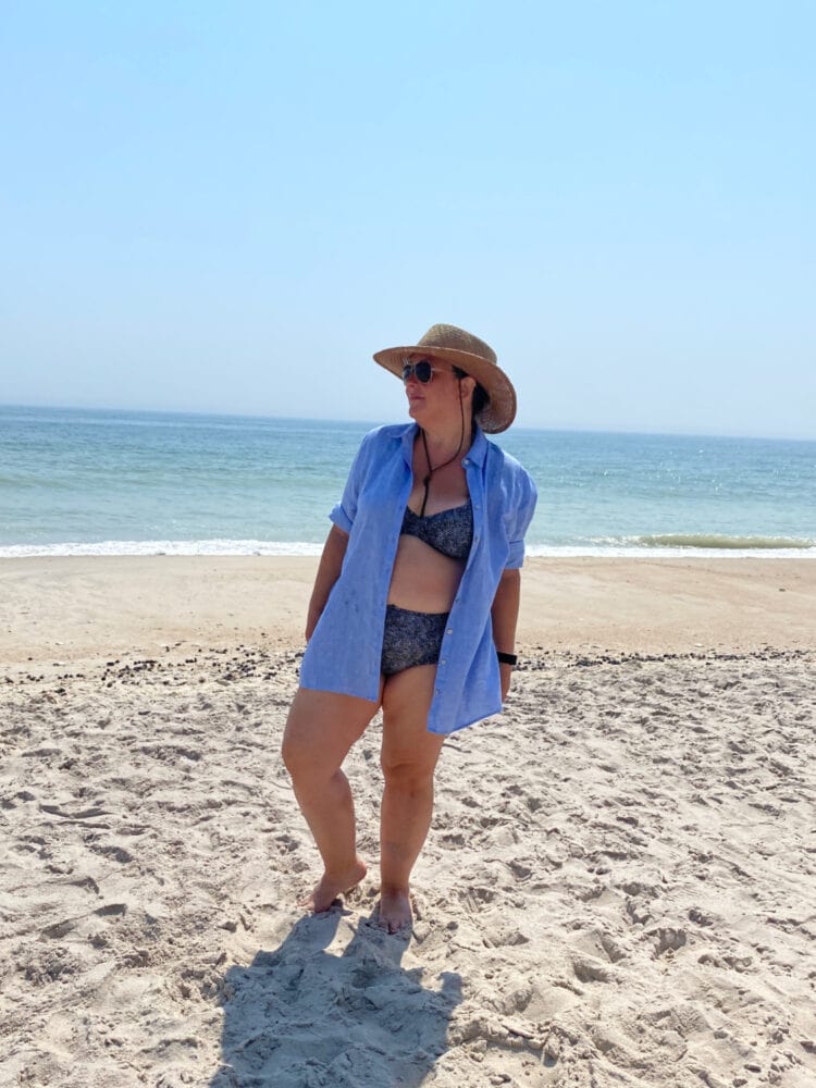 Alison Gary of Wardrobe Oxygen on the beach at Davis Park, Fire Island, wearing a black and white print bikini, blue linen shirt unbuttoned, and a large straw hat
