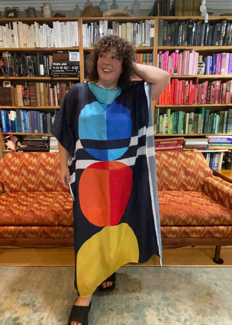Alison Gary of Wardrobe Oxygen standing in her living room in front of an orange upholstered MCM sofa. She is wearing a colorful caftan with black HOKA recovery slides.