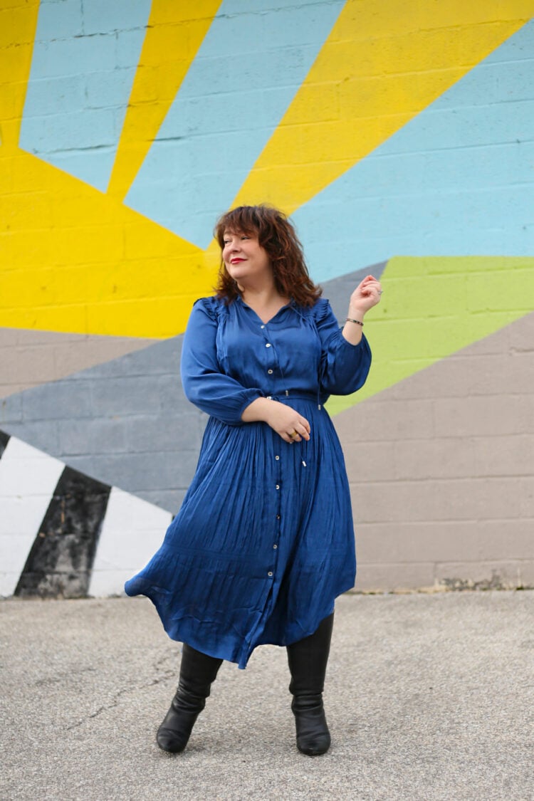 I Look Better In It Than The Model 1 Alison wearing a blue silky drawstring waist longsleeved dress with heeled black knee high boots. She is standing in front of a graphic mural and the wind is catching the hem of her skirt.