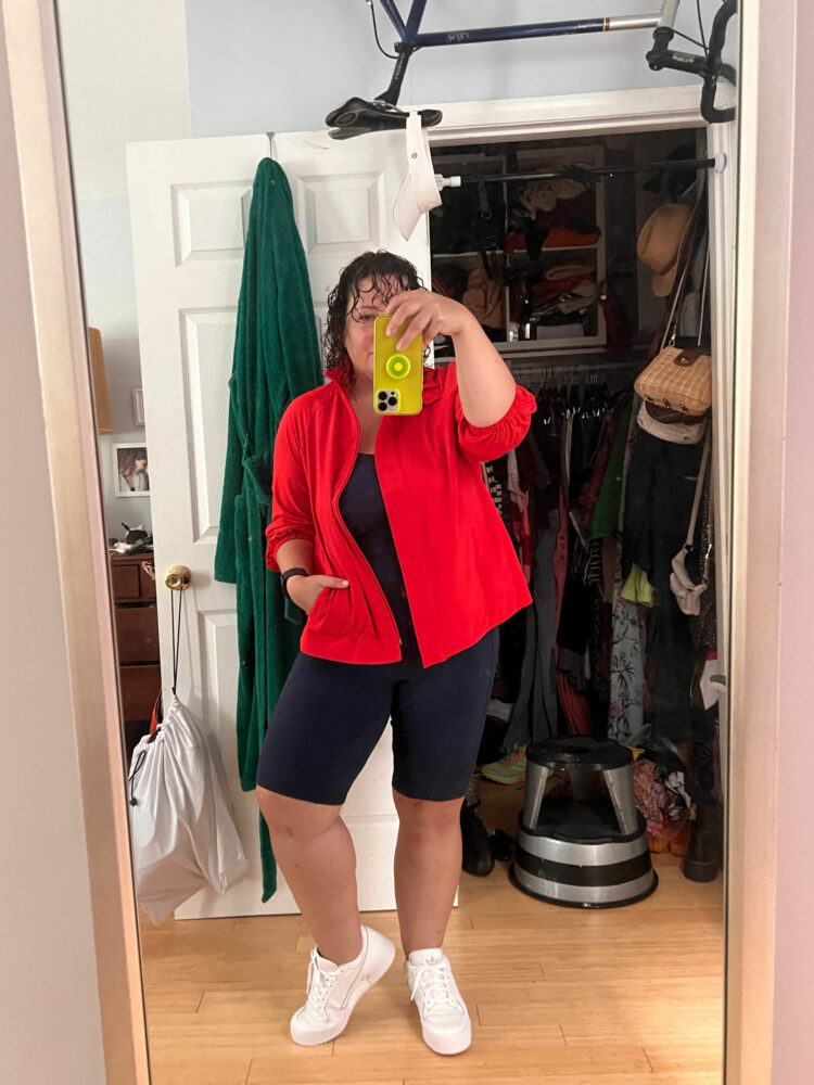 Alison Gary of Wardrobe Oxygen taking a mirror selfie in front of her closet. She is wearing a navy shorts-length unitard and a red windbreaker