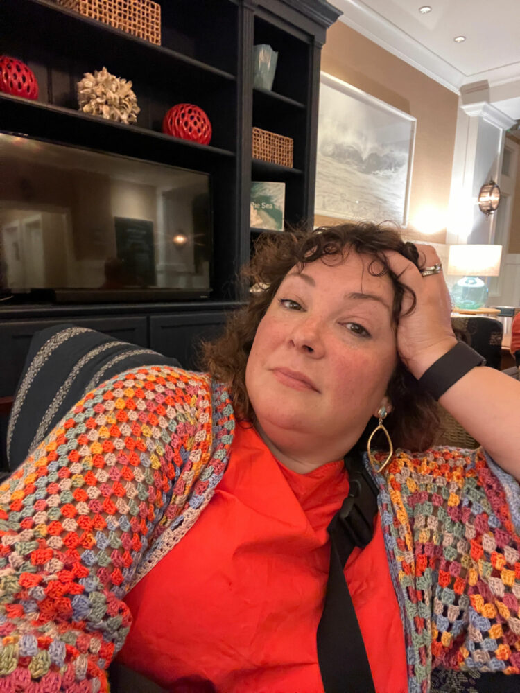 Alison Gary of Wardrobe Oxygen sitting on a sofa wearing an orange cotton sundress and a multicolored crocheted cardigan