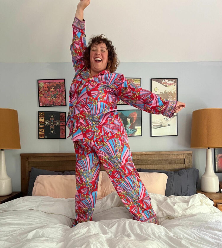 Alison Gary of Wardrobe Oxygen jumping on a bed wearing red printed satin pajamas