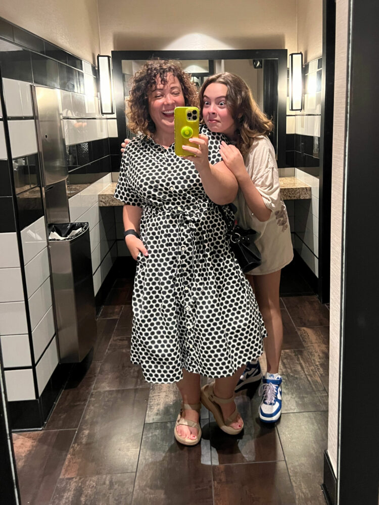 Alison Gary of Wardrobe Oxygen with her daughter in a restaurant bathroom taking a mirror selfie. Alison is wearing a black and white polka dot cotton shirtdress with Teva sandals