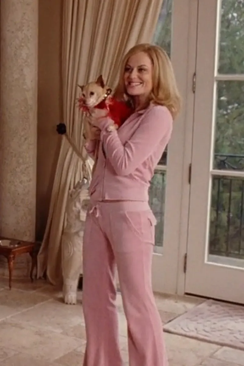 woman in fitted pink sweatsuit with blonde hair. She is holding a tiny dog in her arms; the dog is wearing a red frilly dress.
