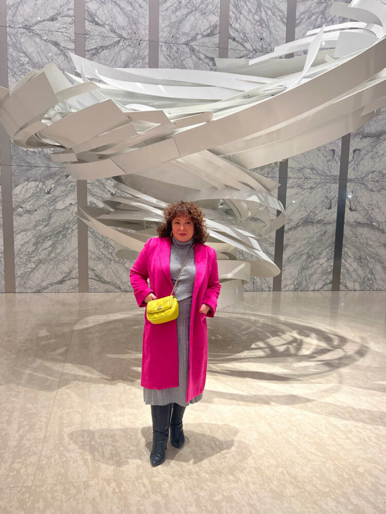 Dressing for the Occasion and the Cold 1 Woman in a gray Quince cashmere turtleneck and matching pleated midi skirt. Over it she is wearing a hot pink wool below knee length coat. She has a pale lime green quilted Coach crossbody over it. On her feet are black knee high boots from Ros Hommerson. She is standing in front of a modern sculpture in the lobby of an office building.