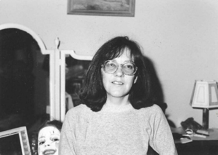 my mom standing in front of her white dressing table, I am peeking out from behind her and my face is painted white with details that makes me think I was a clown or mime
