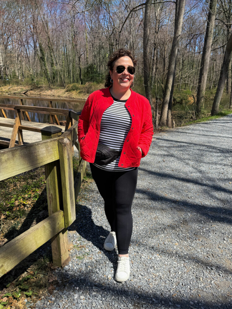 Alison Gary of Wardrobe Oxygen in a red quilted jacket, black and white striped tee, black interlock leggings, and a black belt bag. She is walking on a path in a wooded area with a lake