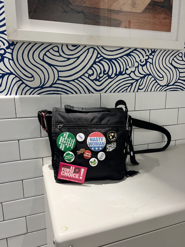 a black nylon canvas crossbody bag sitting on a table. It is covered with buttons regarding social issues like abortion and women's rights