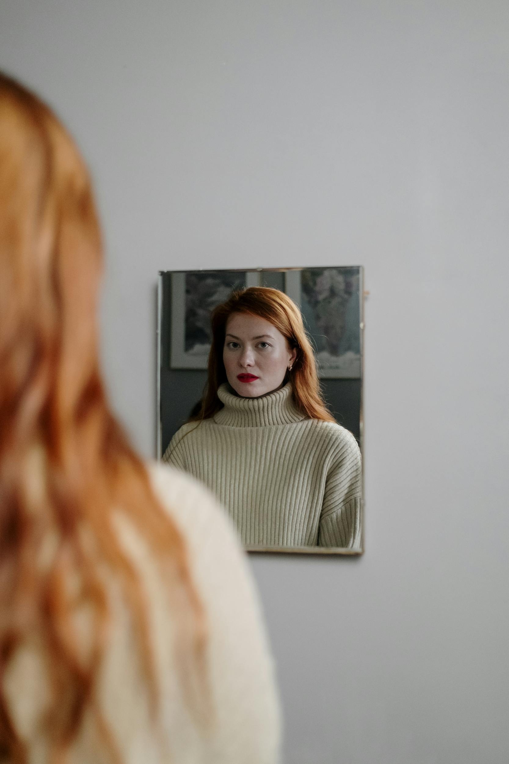 A redhead woman gazes pensively at her reflection in a mirror, wearing a knitted turtleneck indoors.