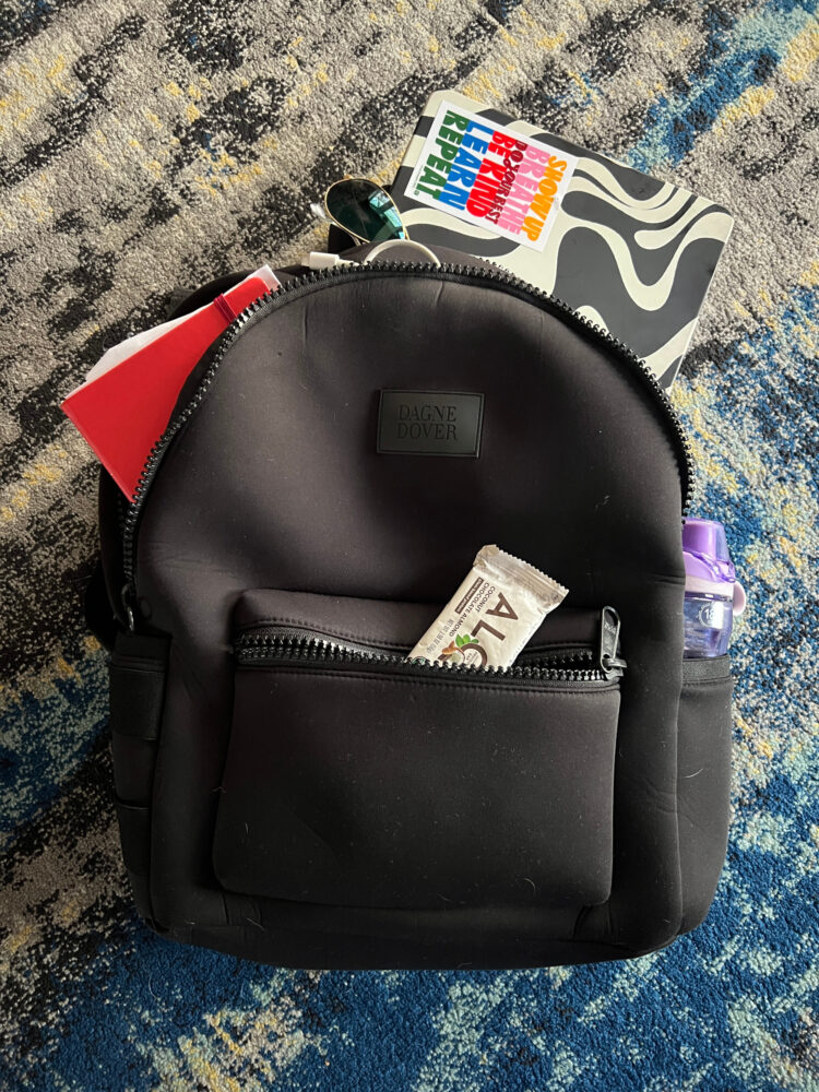 dagne dover large dakota backpack review showing the black version packed for work with a laptop, notebook, water bottle, charging cords, and a protein bar.