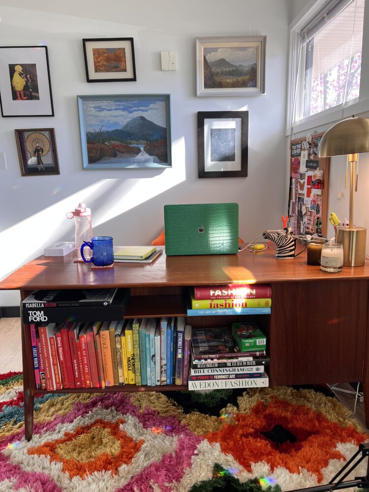 The Business Side of Content Creation: What I've Learned the Hard Way 3 Alison Gary's office for Wardrobe Oxygen at the Granite Building in Greenbelt, Maryland. Photo shows a mid-century modern desk in an office with a laptop o top. There is a colorful shag carpet and a shelf in the desk is full of colorful books about art and fashion. A gallery wall behind the desk holds framed paintings and prints including one of Diane von Furstenberg with Big Bird which was from a Bazaar magazine photo shoot.
