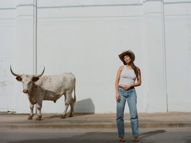 Weekend Reads for March 14 & 15 10 kacey musgraves in jeans and a white tank with cowboy hat standing in front of a white wall with a steer in the background